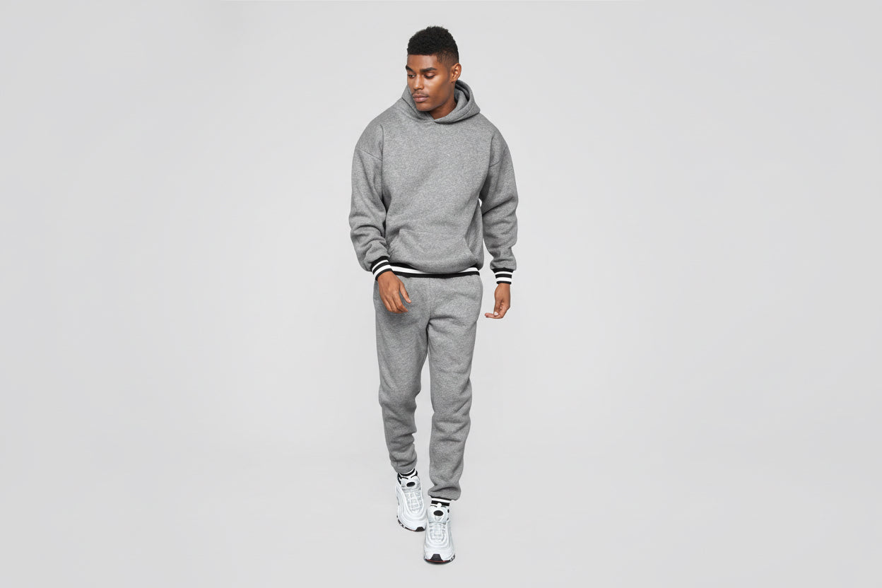 Man wearing a gray hoodie and matching sweatpants with white sneakers on a plain background