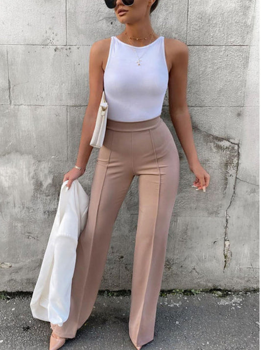 Woman in white sleeveless top, beige high-waisted wide-leg pants, sunglasses, holding white blazer against concrete wall