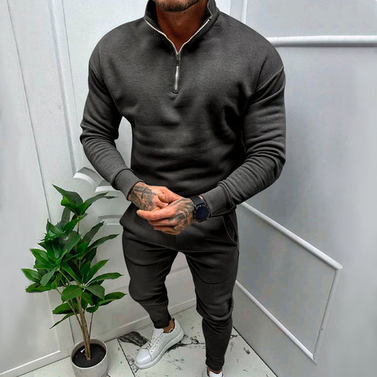 Man wearing dark gray zip-up sweatshirt and matching joggers with white sneakers, standing indoors near green potted plant