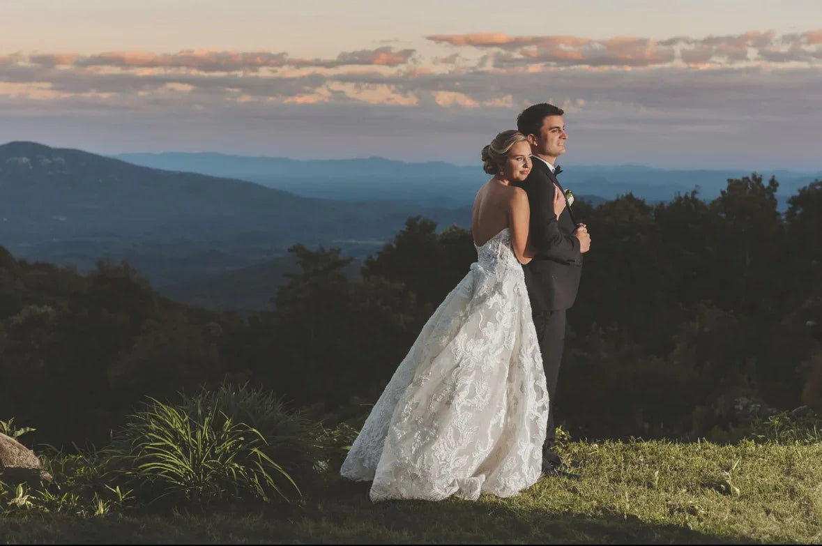 Bride in lace wedding dress hugging groom in black suit, scenic mountain landscape at sunset