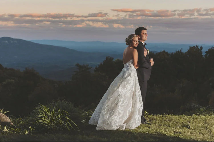 Bride in lace wedding dress hugging groom in black suit, scenic mountain landscape at sunset