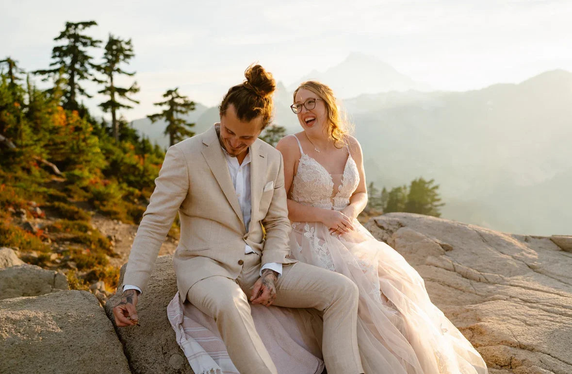 Smiling bride in lace dress and groom in beige suit sitting on rocky mountain with forest backdrop