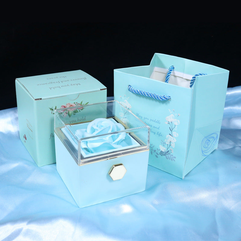 Blue gift box with clear lid and artificial blue rose, next to matching gift bag and floral box