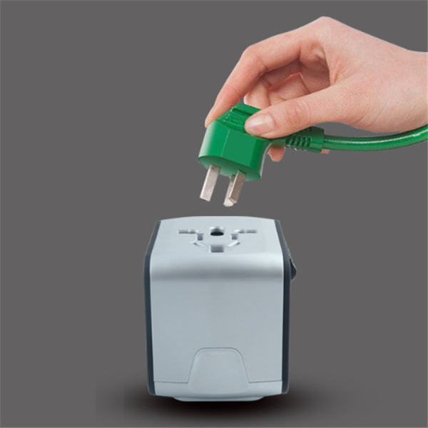 Hand holding green electrical plug above silver universal power adapter socket on gray background
