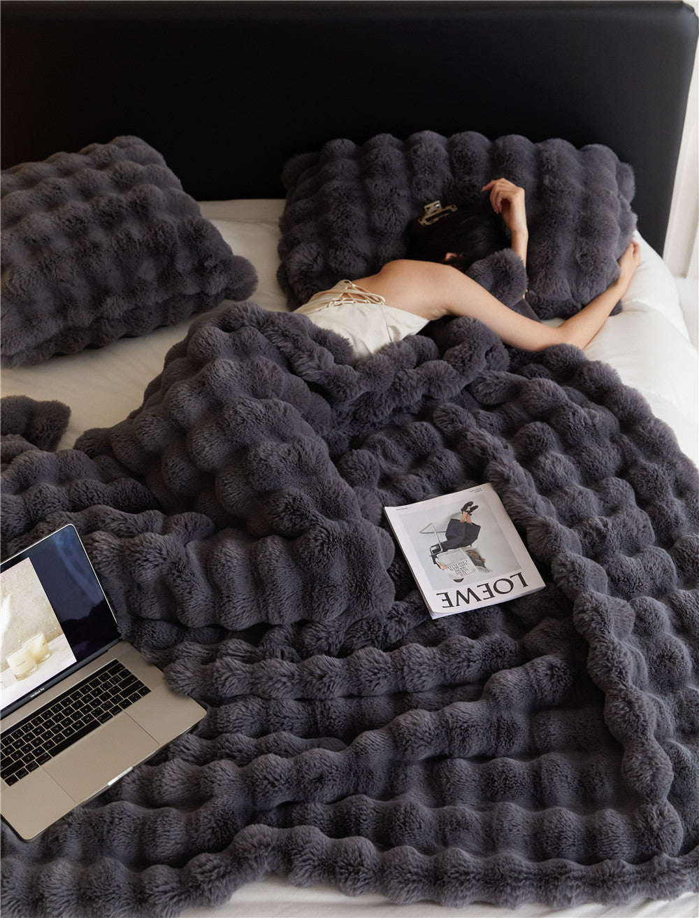 Woman resting on bed with luxurious dark gray fur blanket and matching pillows, laptop and magazine nearby