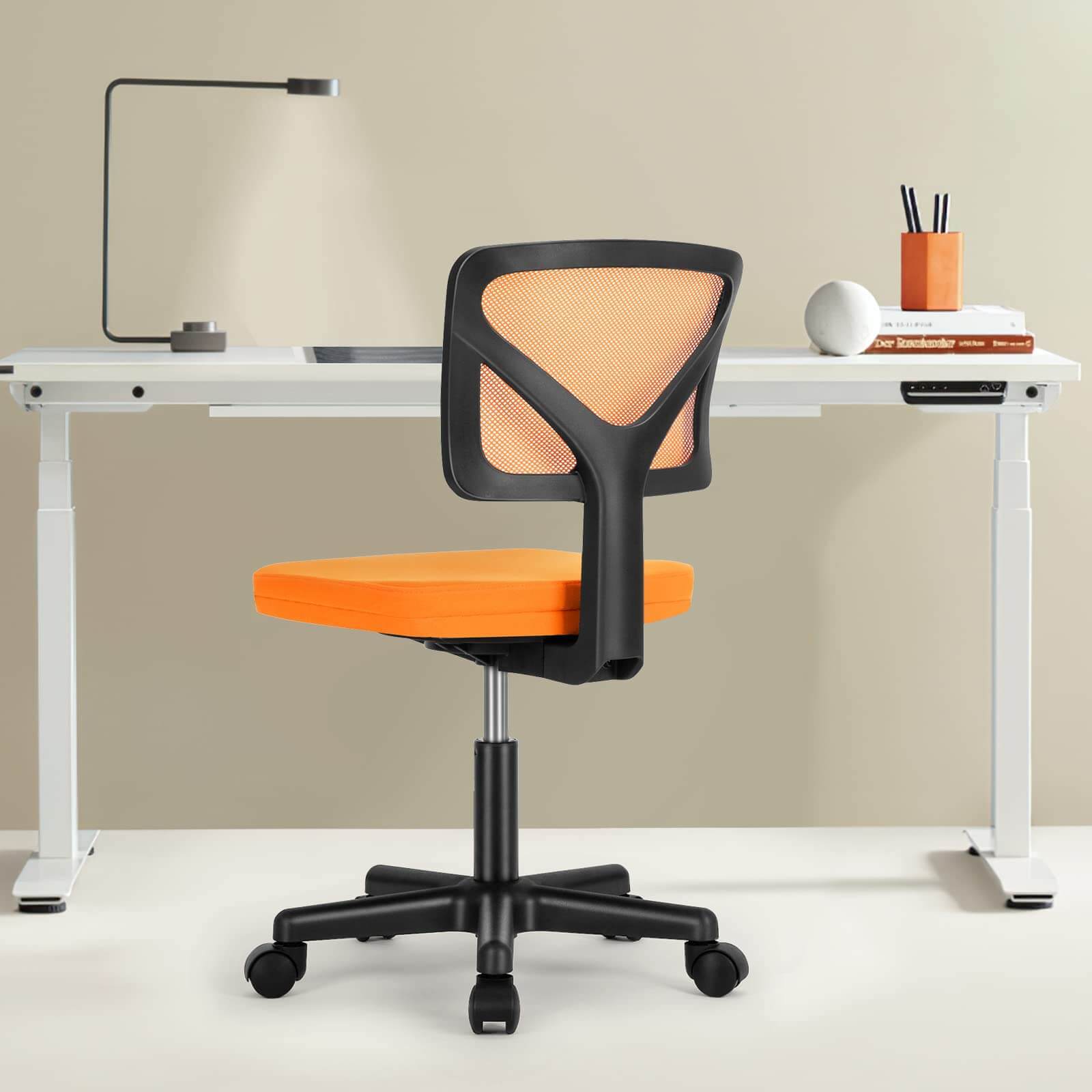 Modern office chair with orange mesh back and seat in front of white desk with lamp and books