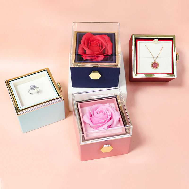Luxury jewelry boxes with clear lids displaying a diamond ring, pink pendant necklace, red rose, and pink rose on pastel background