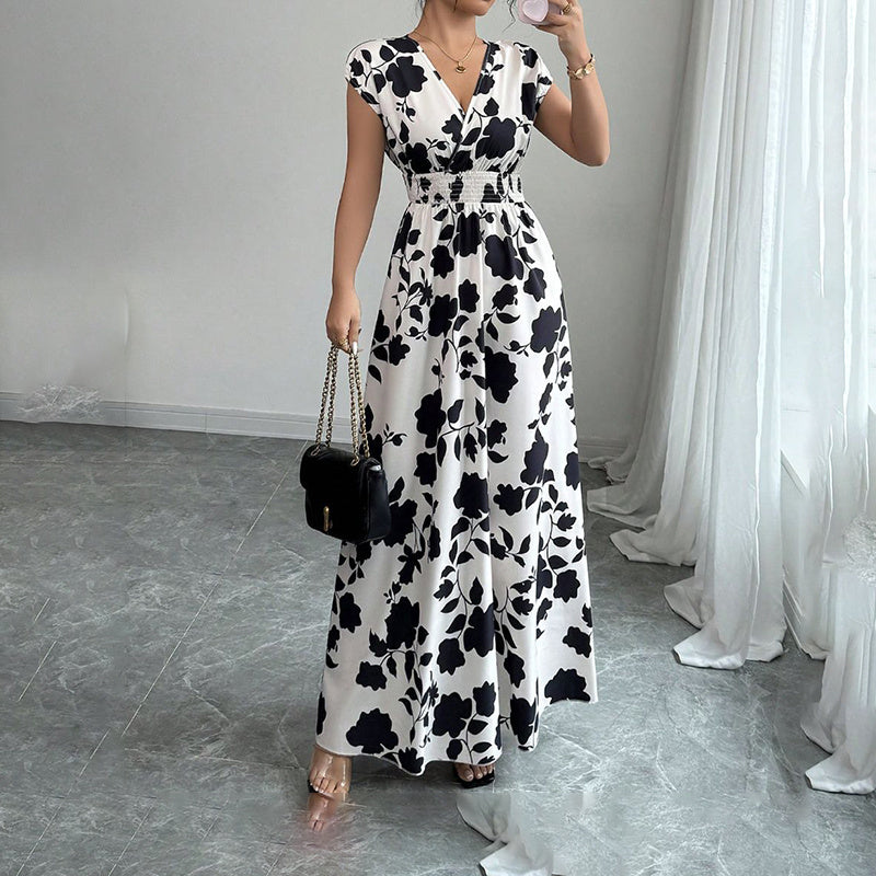 Woman in sleeveless white maxi dress with black floral print, holding black handbag, standing on gray floor