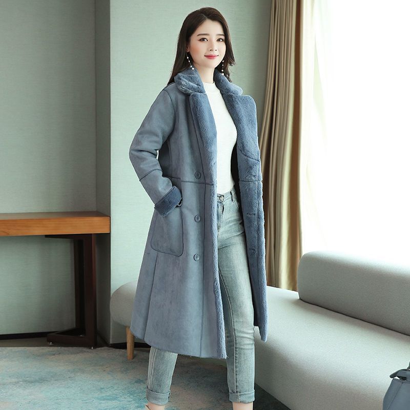 Woman wearing long blue fur-lined winter coat with pockets and cuff details, standing indoors