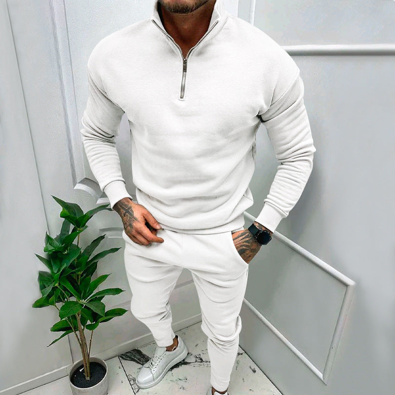 Man wearing white zip-up sweatshirt, matching joggers, and white sneakers in minimalist room with plant