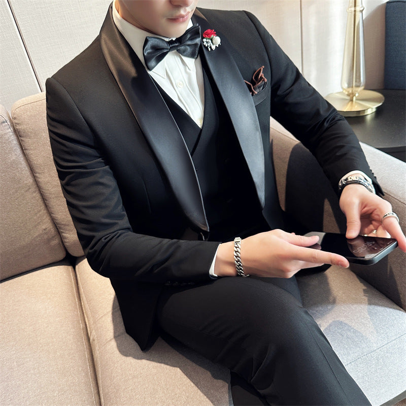 Man in black tuxedo with satin lapels, white shirt, black bow tie, floral lapel pin, and accessories sitting on beige sofa using smartphone