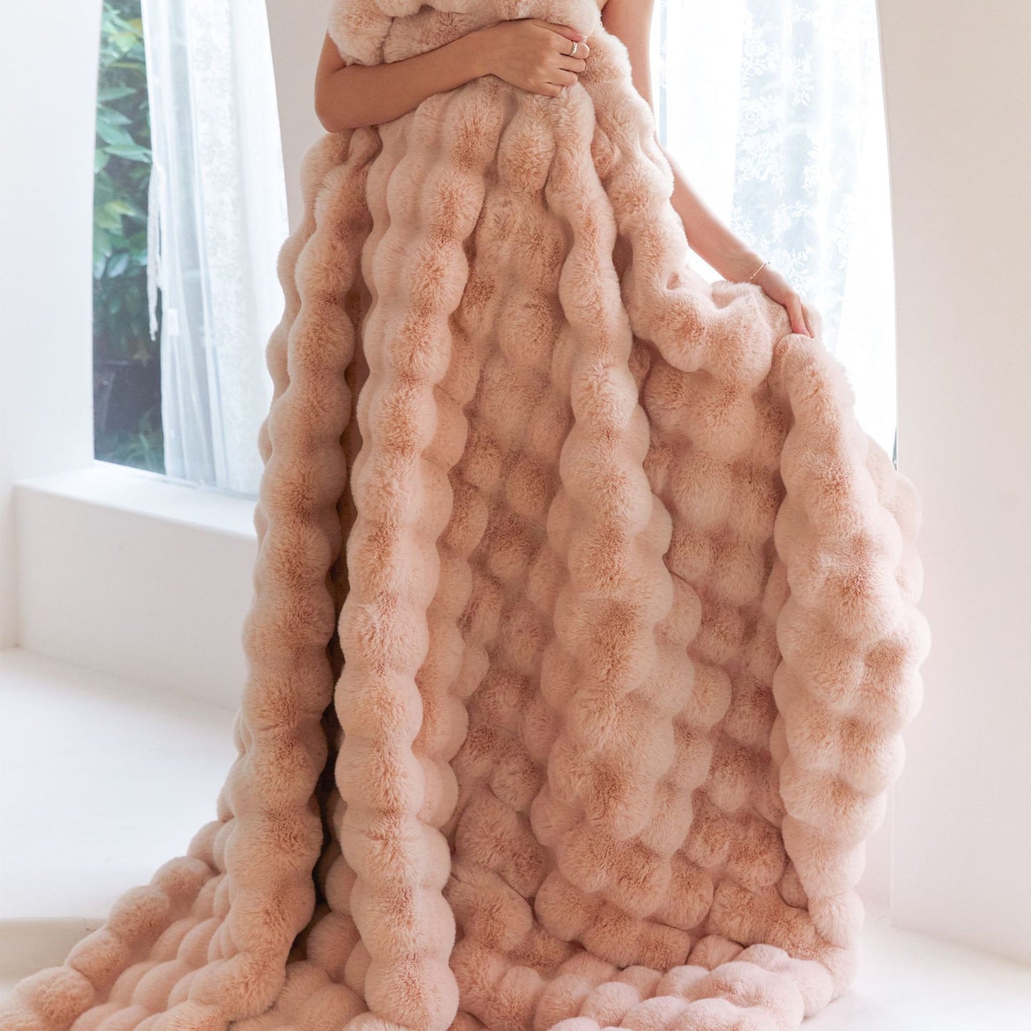 Person holding a soft, plush, blush pink textured blanket indoors near a window with lace curtains