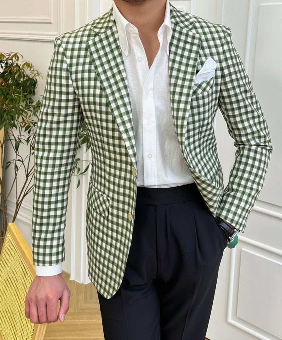 Man wearing green and white checkered blazer, white shirt, black pants, standing indoors by a chair
