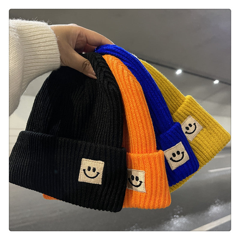 Hand holding colorful ribbed knit beanies in black, orange, blue, and yellow with smiley face patches