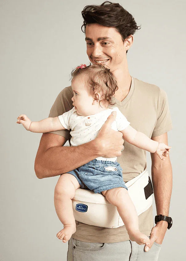 Smiling man carrying baby in beige baby hip seat carrier with blue denim shorts