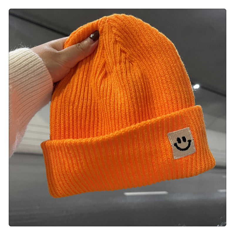 Hand holding bright orange ribbed knit beanie with cuff and smiley face patch