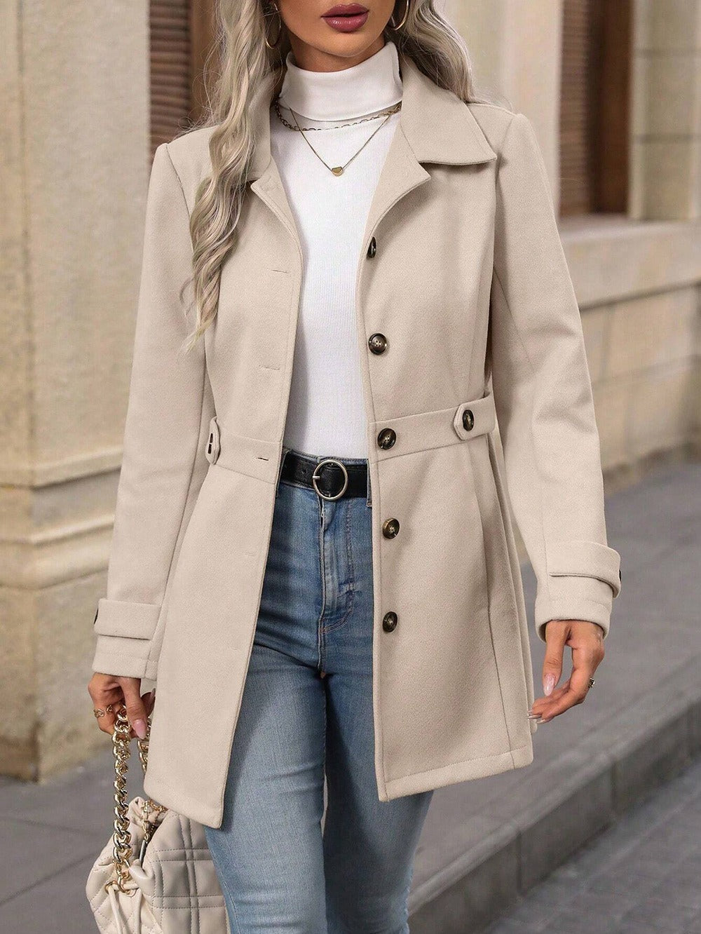 Woman wearing beige buttoned coat, white turtleneck, blue jeans, and carrying quilted handbag in urban setting
