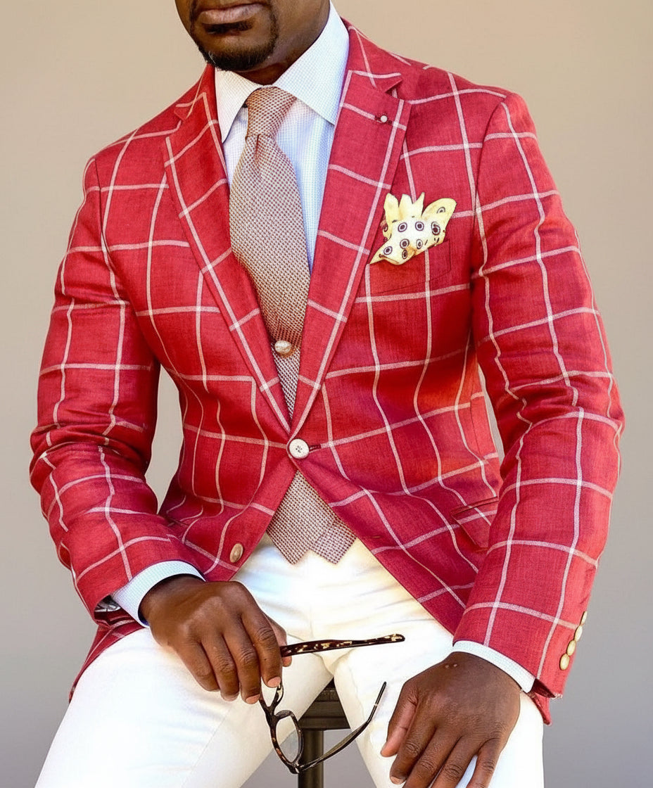 Man wearing red checkered blazer with light brown tie, beige pocket square, white pants, holding glasses