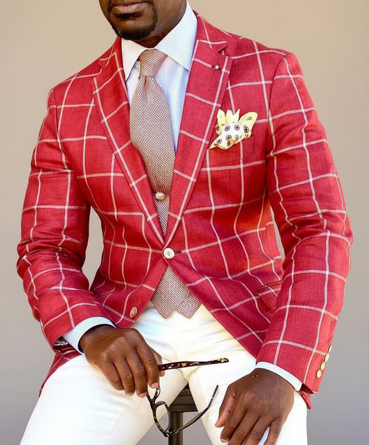 Man wearing red checkered blazer with light brown tie, beige pocket square, white pants, holding glasses