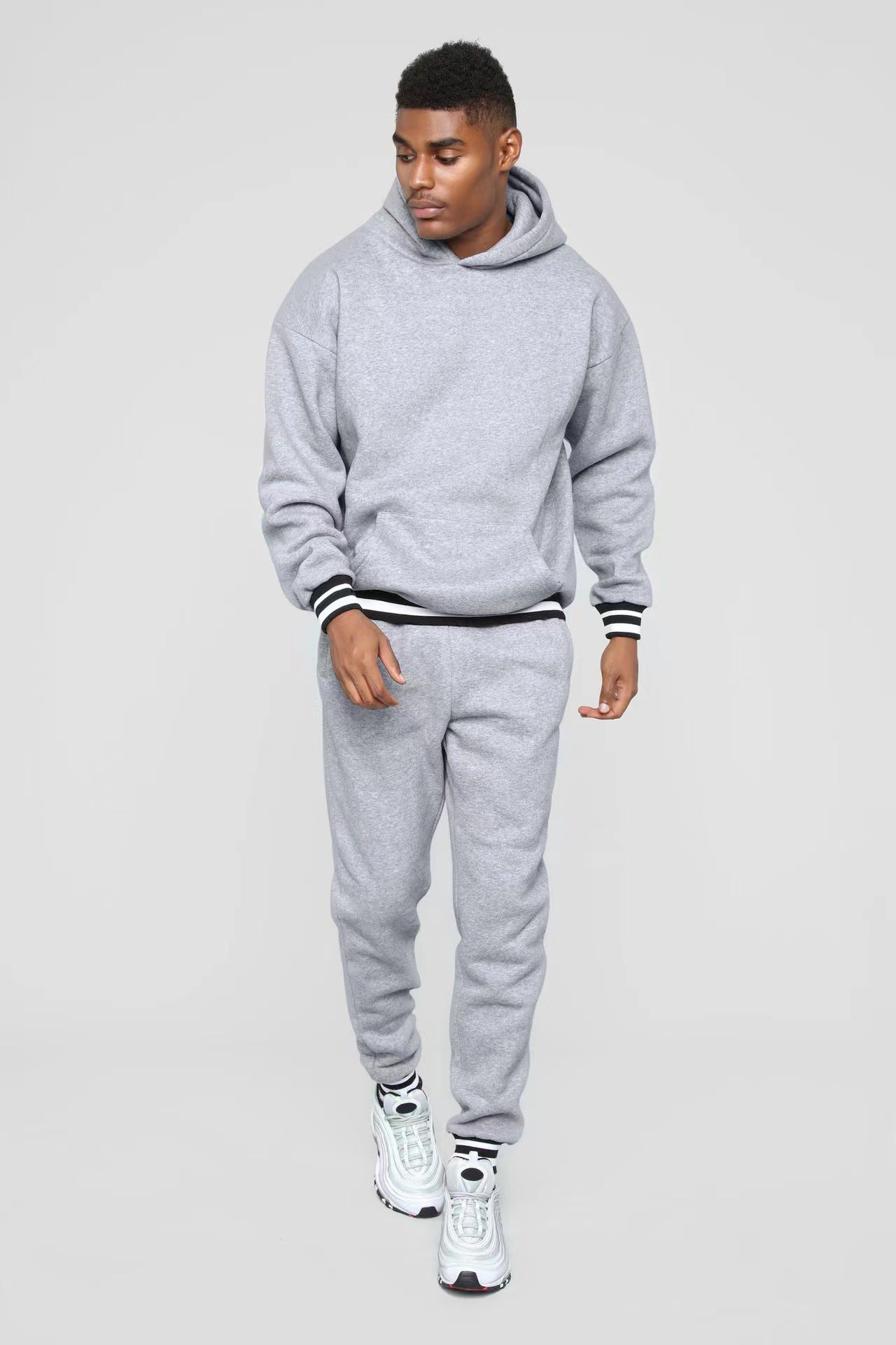 Young man wearing a gray hoodie and matching sweatpants with white sneakers on gray background