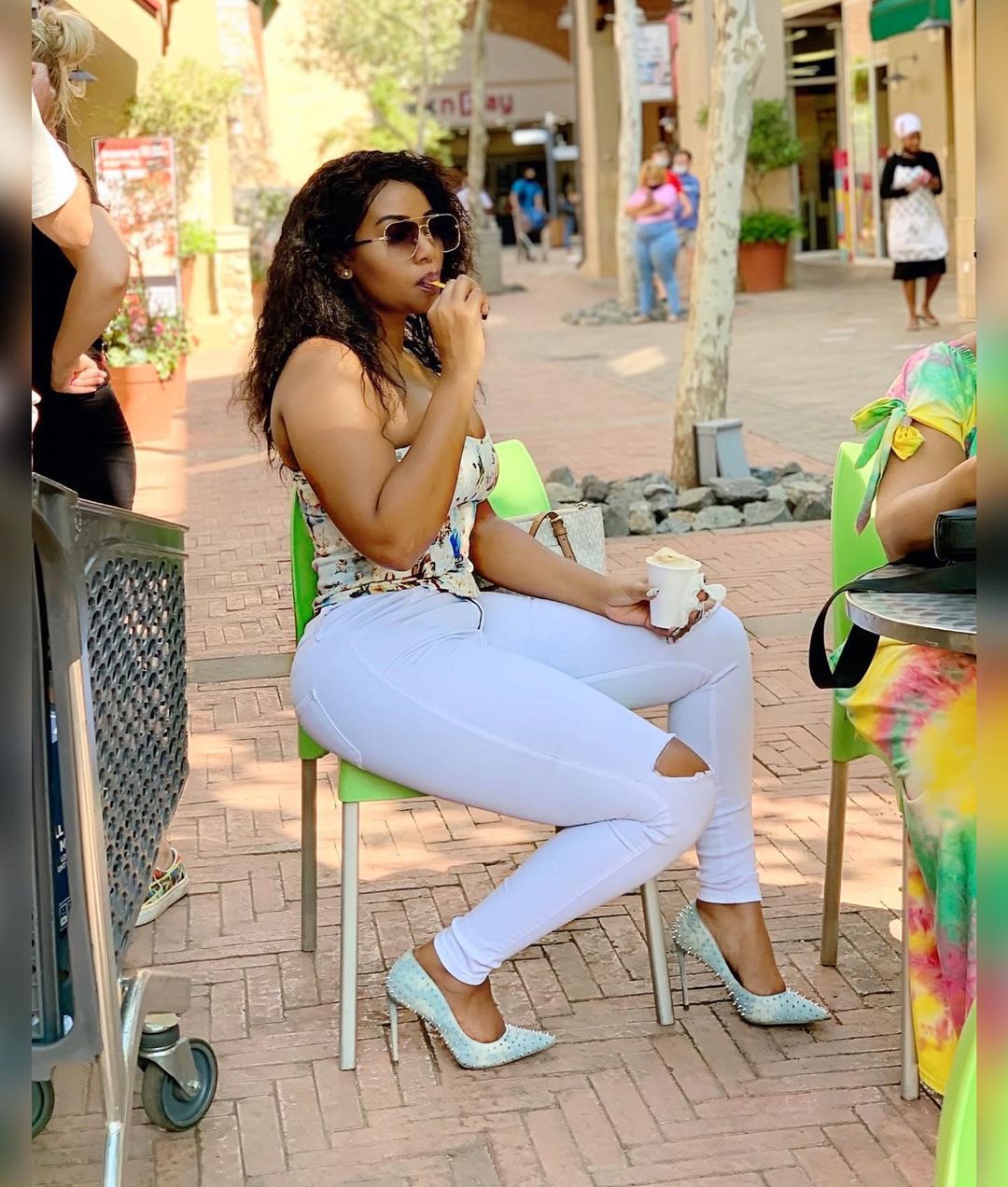Stylish woman wearing white ripped jeans and studded heels sitting outdoors, enjoying coffee and a snack