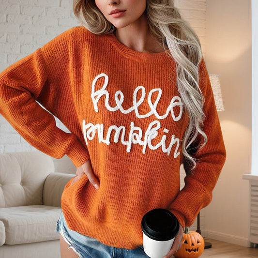 Woman wearing orange sweater with white "hello pumpkin" text, holding coffee cup in cozy living room with pumpkin decor