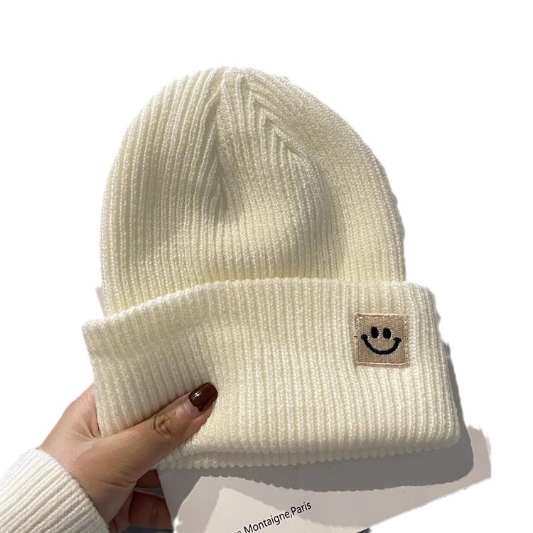 Hand holding cream ribbed knit beanie with folded cuff and smiley face patch