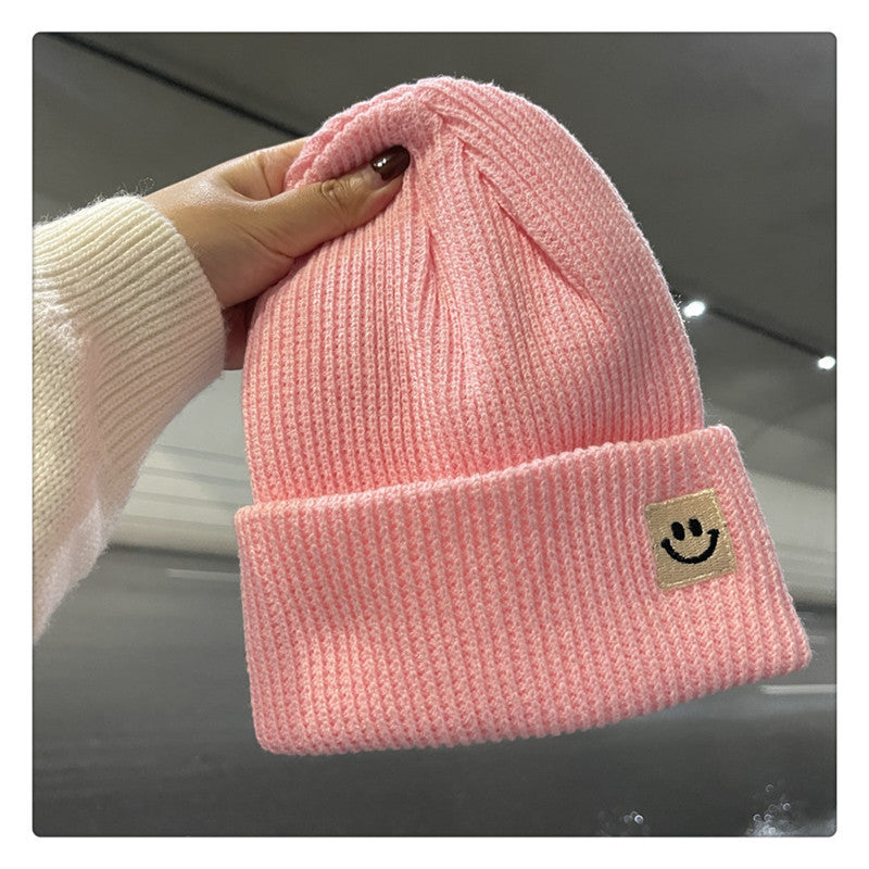 Hand holding a pink knitted beanie hat with a smiley face patch on the cuff