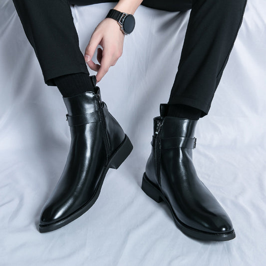 Man wearing black leather ankle boots with side zipper and buckle, black pants, sitting on white surface