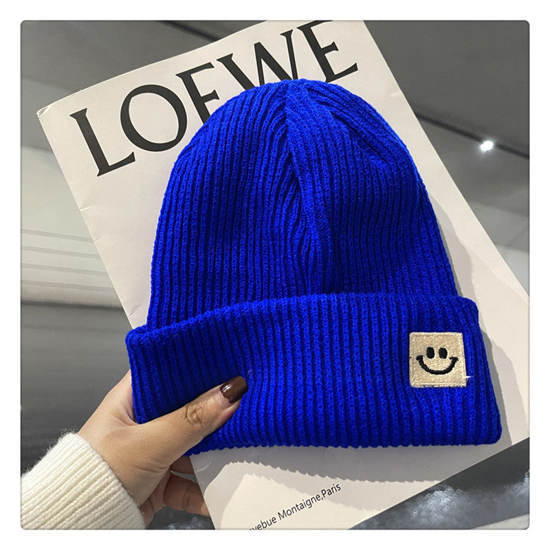 Hand holding bright blue ribbed knit beanie with smiley face patch over Loewe magazine cover