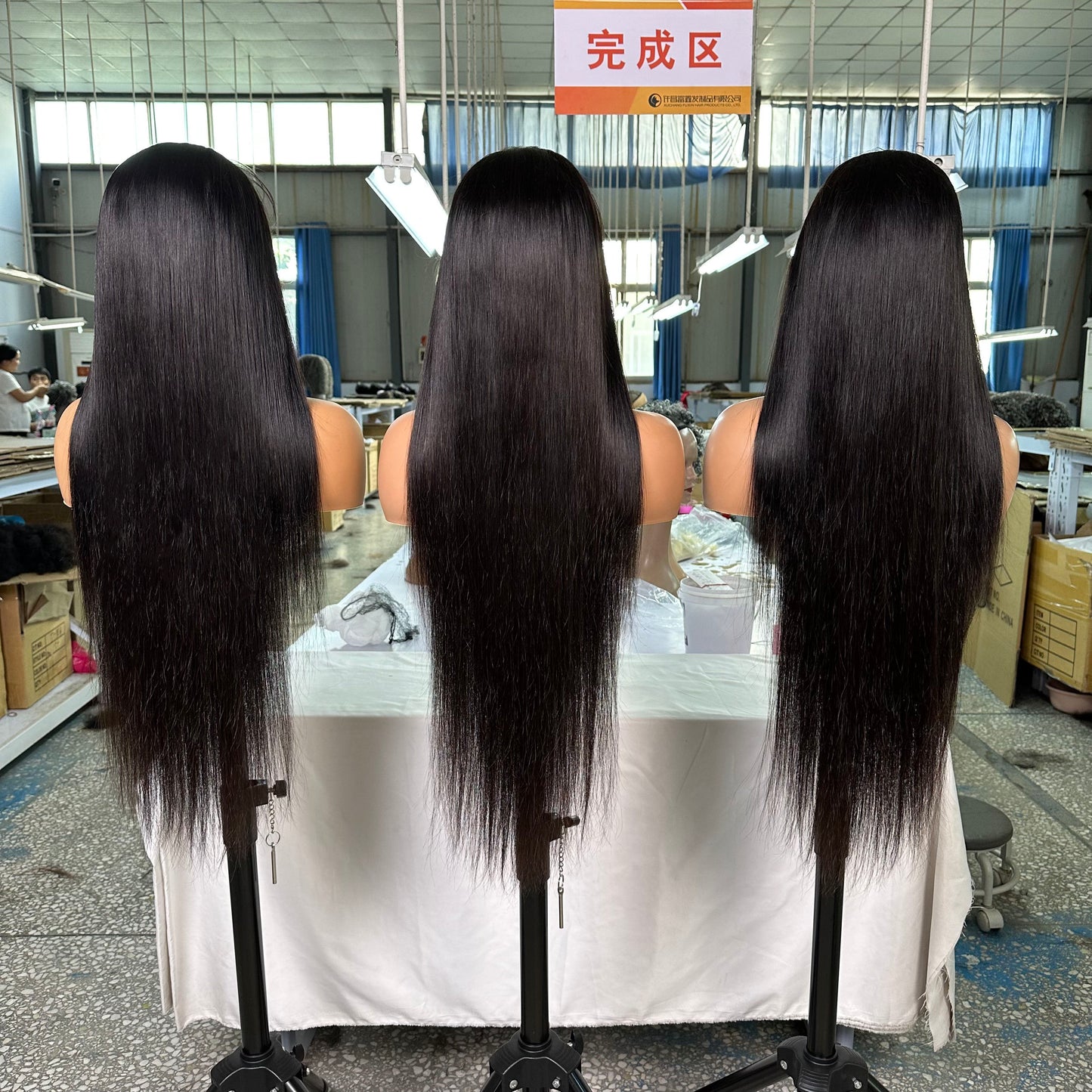 Three long black straight hair wigs displayed on mannequin heads in a brightly lit wig factory