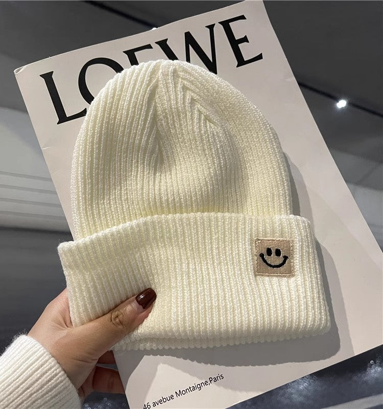 Hand holding white ribbed knit beanie with smiley face patch over LOEWE magazine cover