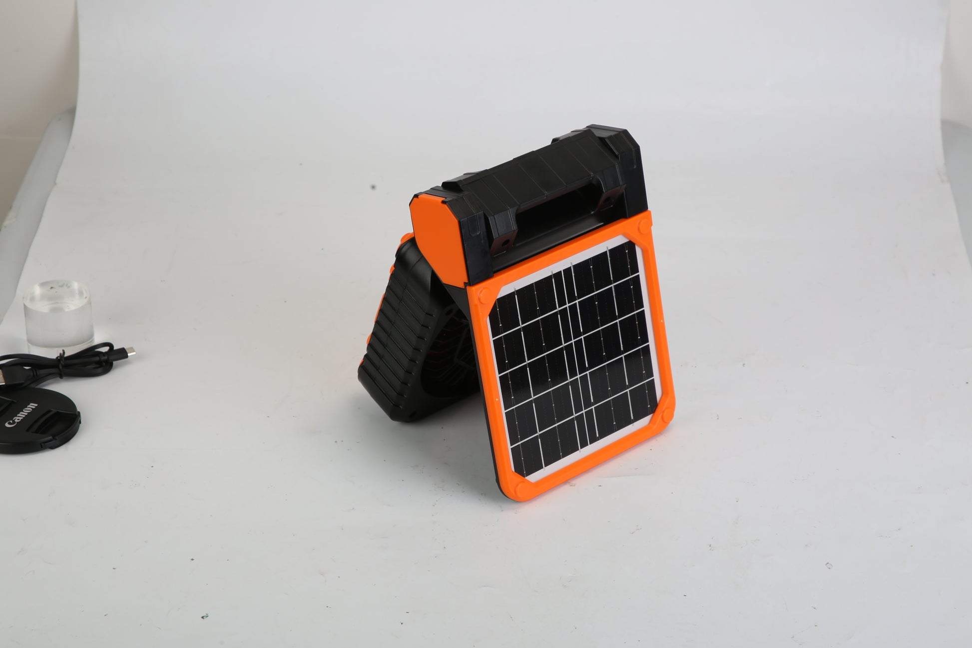 Portable black and orange solar panel device with handle on white background, USB cable and Canon lens cap nearby