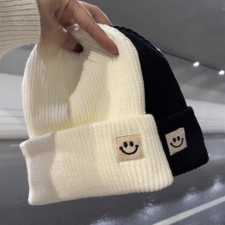 Hand holding white and black ribbed knit beanies with smiley face patches in indoor setting