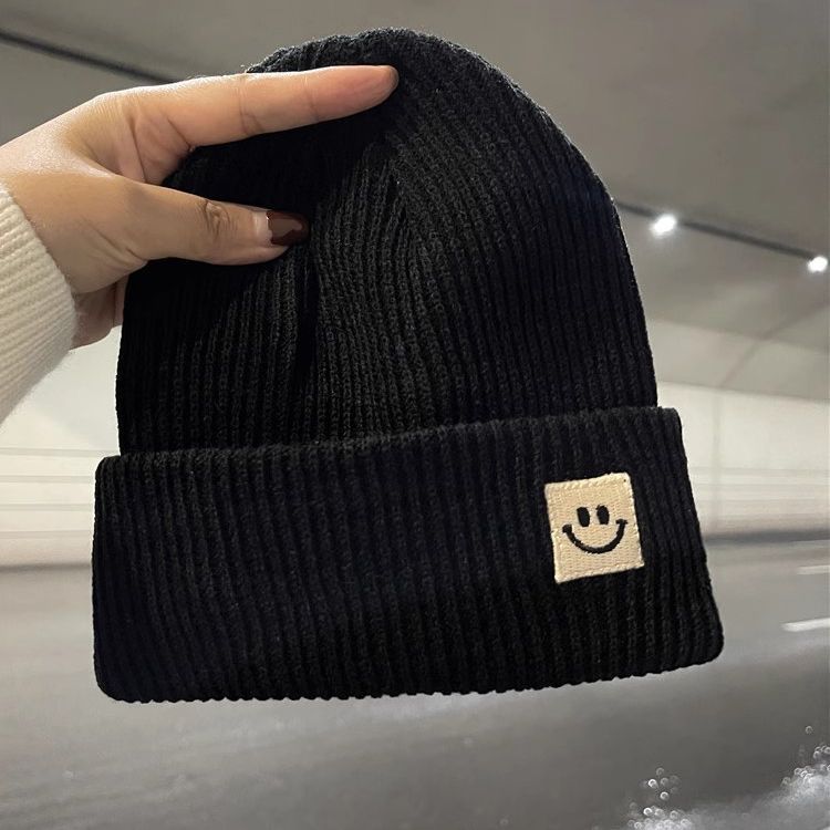 Hand holding a black ribbed knit beanie with a beige smiley face patch in a blurred indoor setting