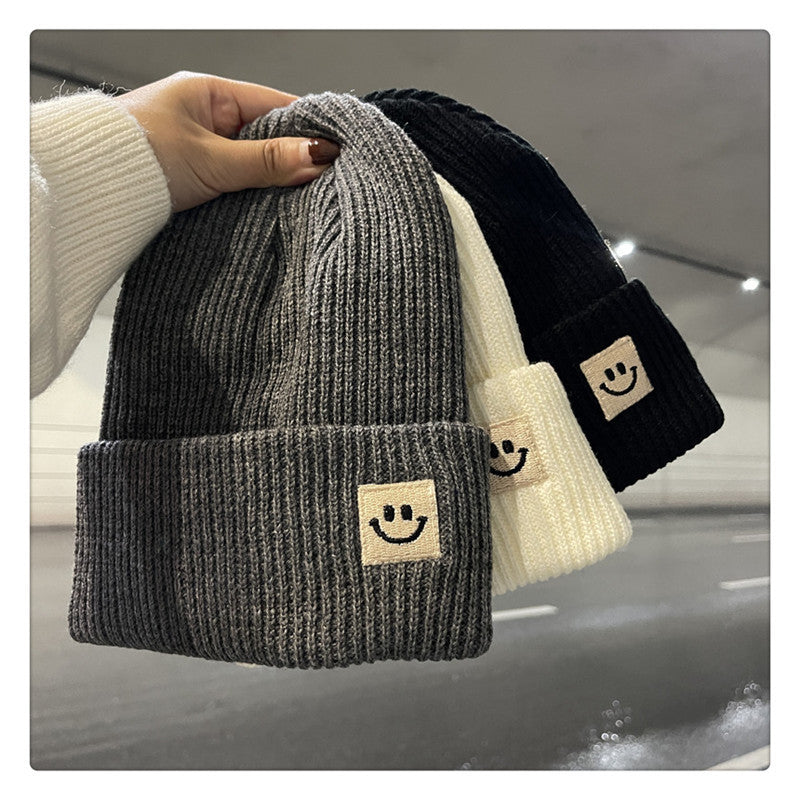 Hand holding three ribbed knit beanies in grey, white, and black with smiling face patches