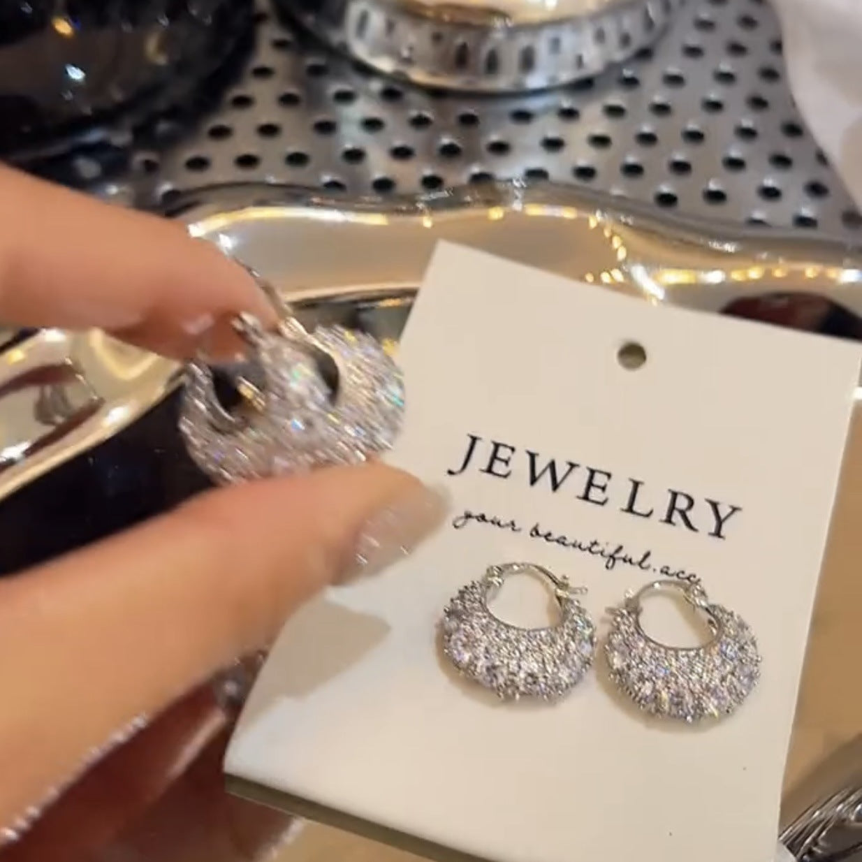 Sparkling silver hoop earrings with crystals on white jewelry card held by hand on reflective metal tray