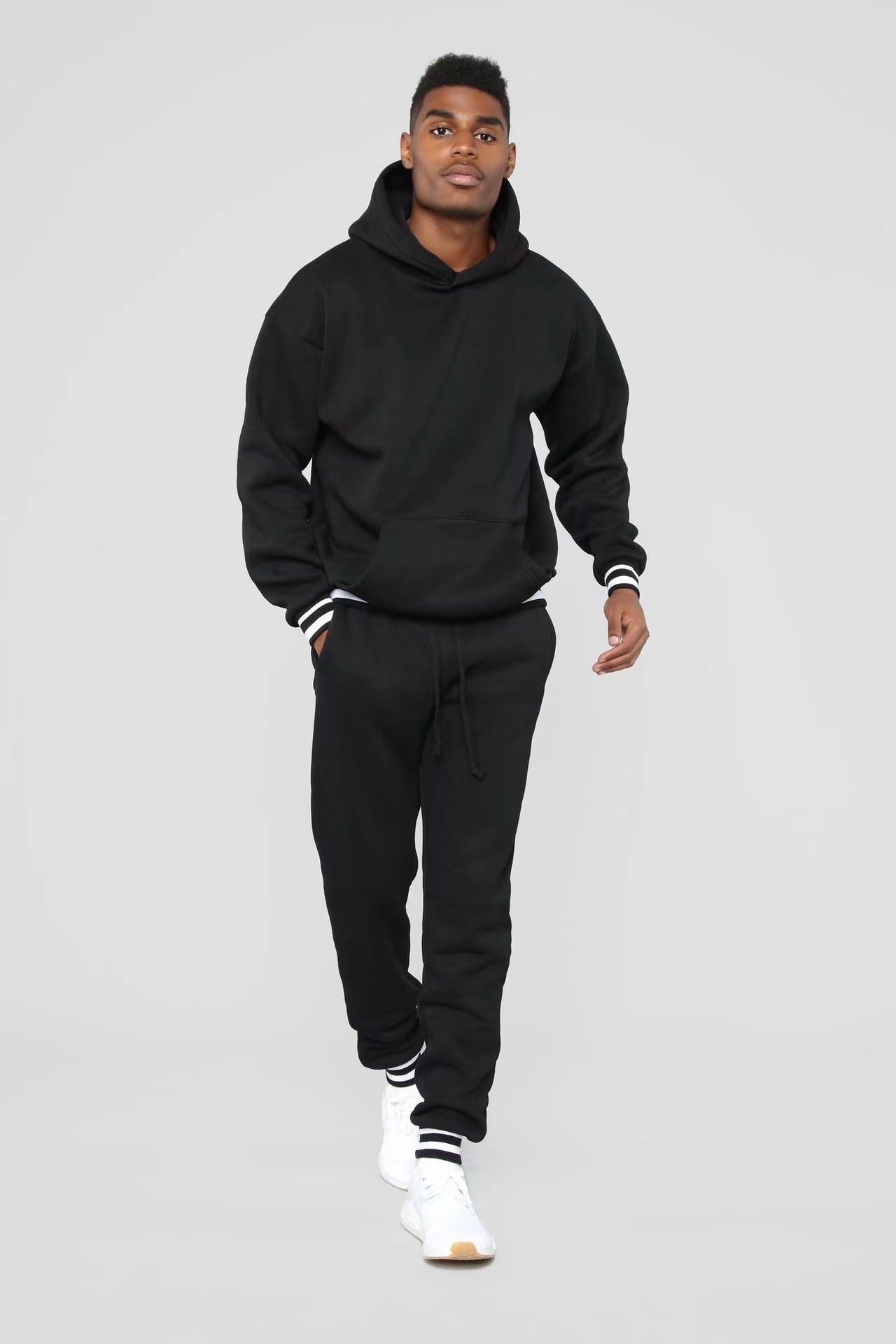 Young man wearing black hoodie and matching joggers with white sneakers walking on gray background