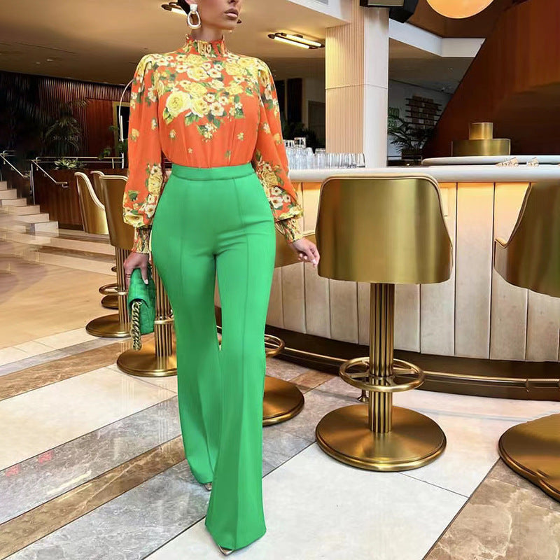 Woman in an orange floral blouse and bright green high-waist pants holding a green clutch in a modern bar