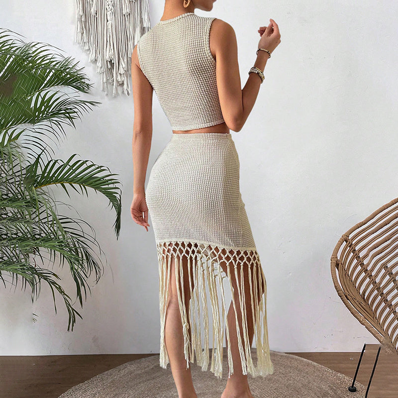 Woman in sleeveless beige knit top and fringe hem skirt posing indoors by plant and wicker chair