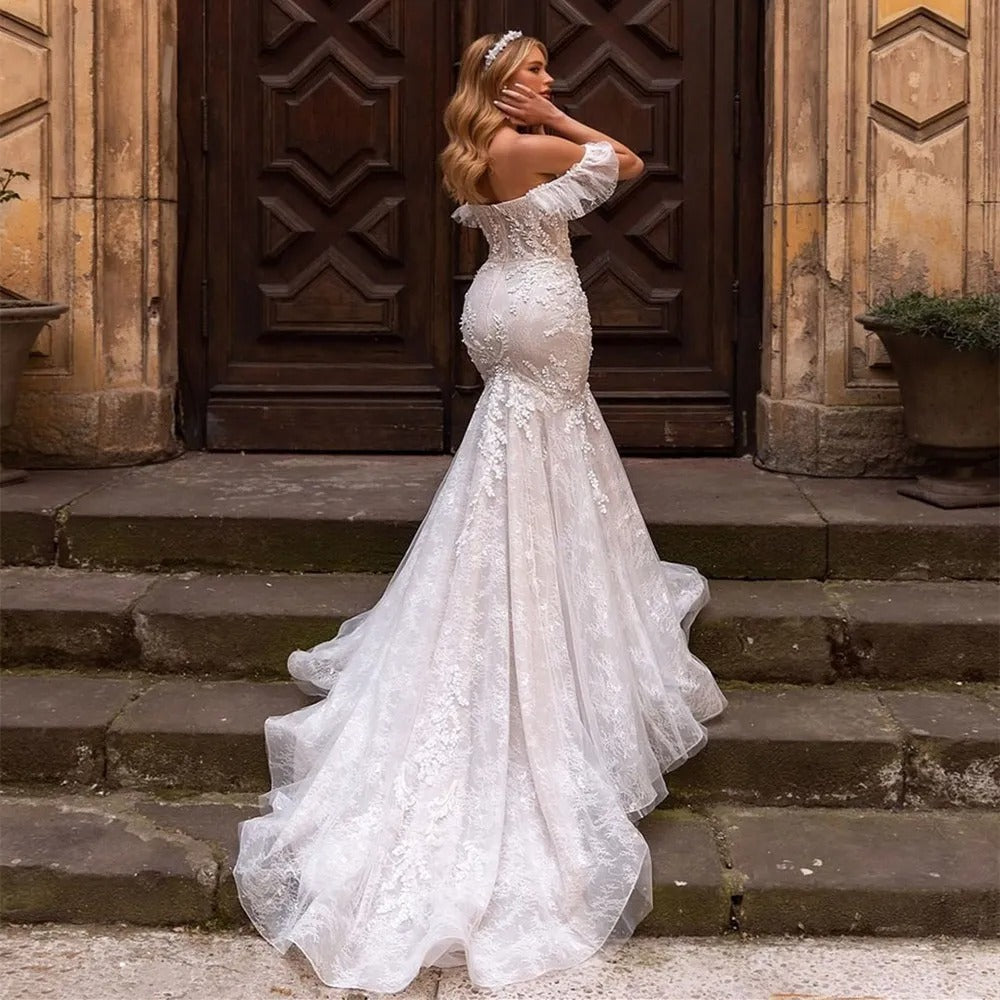 Bride in a lace off-shoulder mermaid wedding dress with a long train, standing on stone steps