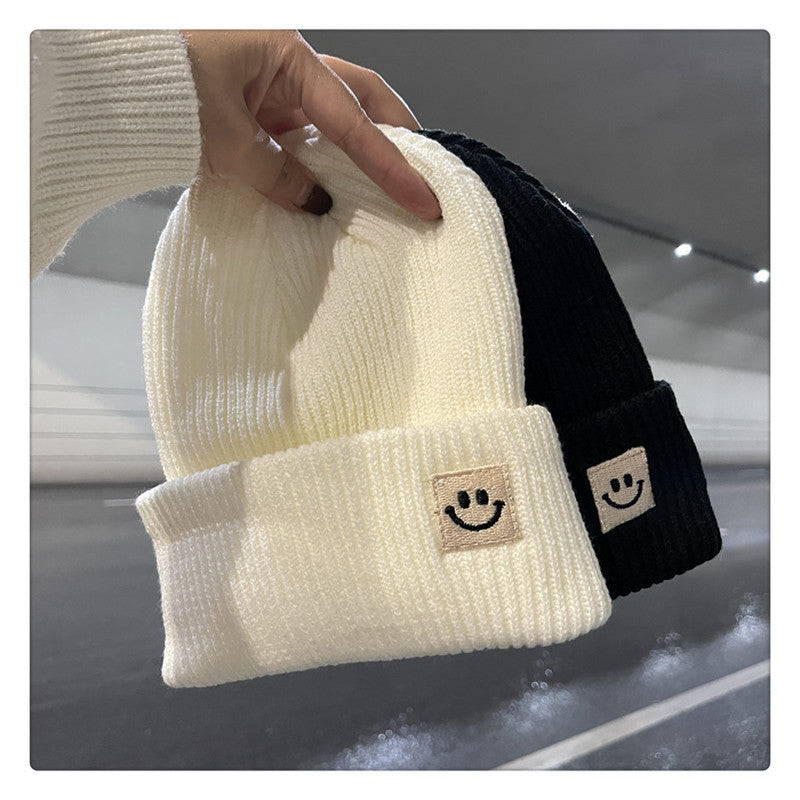 Hand holding white and black ribbed knit beanies with cuff smiley face patches