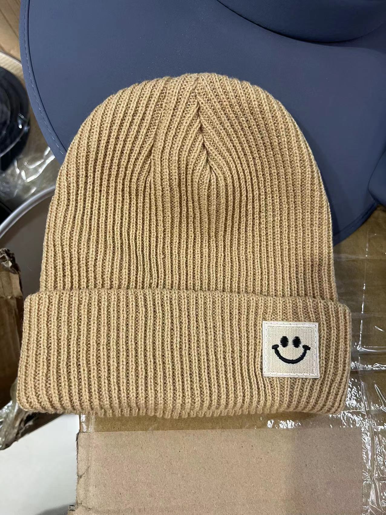 Beige ribbed knit beanie with smiley face patch, placed on cardboard and blue surface