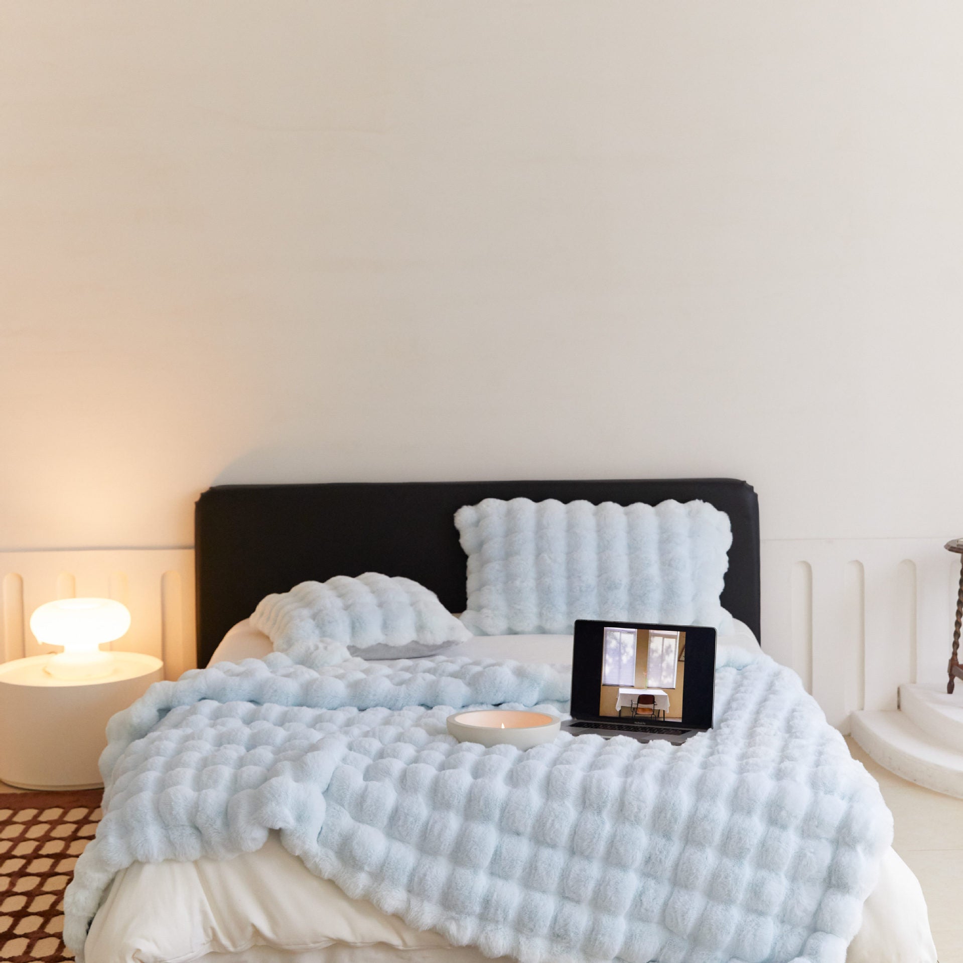 Cozy bedroom with black headboard, textured light blue blanket and pillows, laptop on bed, modern lamp on side table