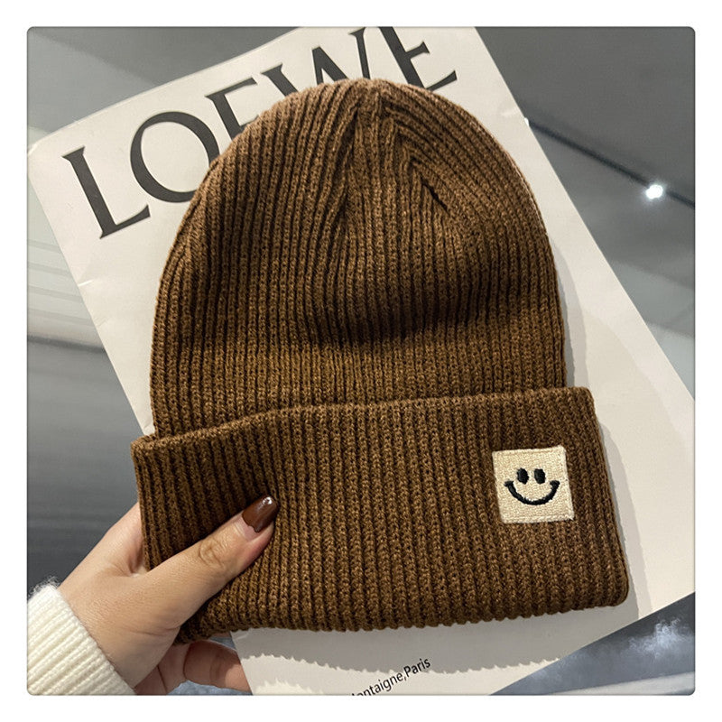 Brown ribbed knit beanie with a small smiley face patch on cuff held over a Loewe magazine