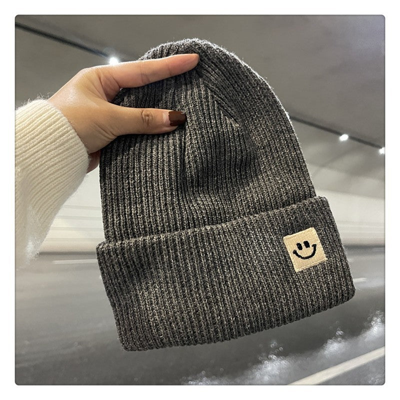 Hand holding dark gray ribbed knit beanie with smiley face patch in dimly lit tunnel
