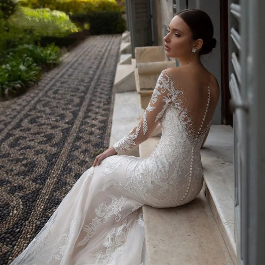 Bride in elegant lace wedding gown with long sleeves and sheer back sitting on stone ledge outdoors