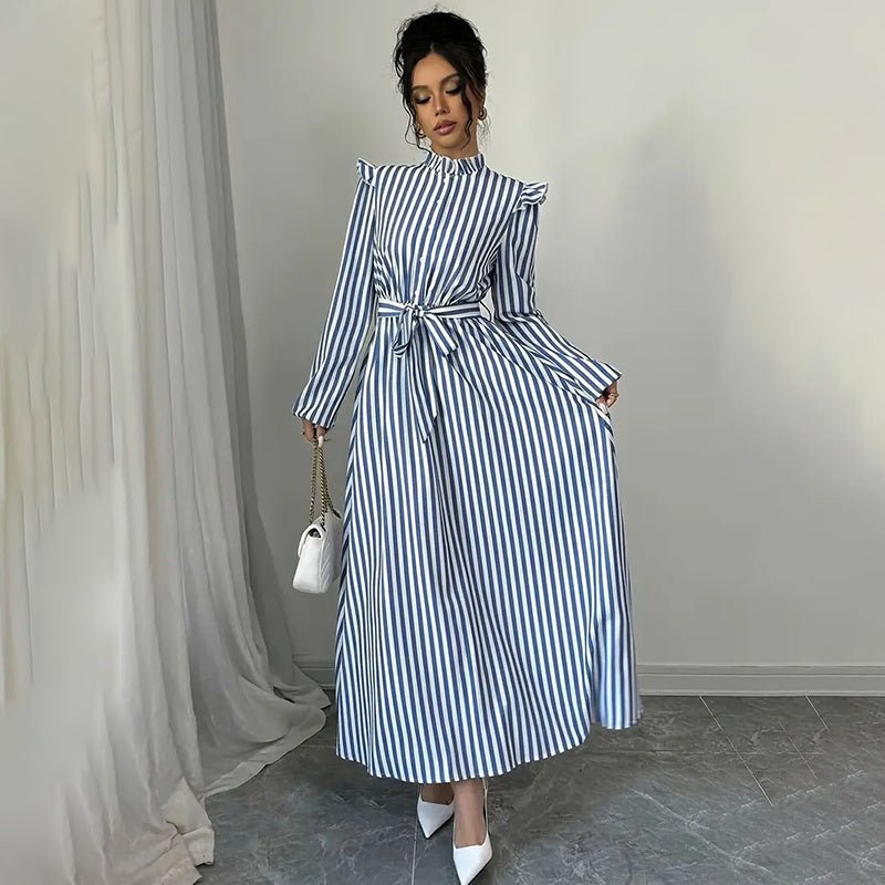 Woman wearing blue and white striped long sleeve maxi dress with stand collar and waist tie, holding white handbag