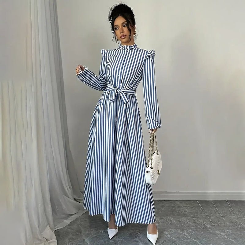 Woman wearing blue and white vertical striped long sleeve maxi dress with stand collar and waist tie, holding white handbag